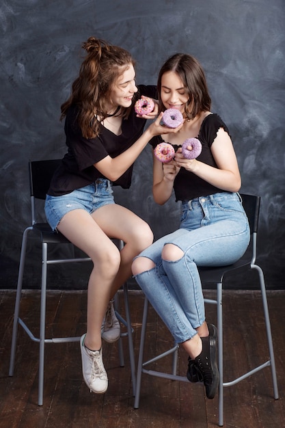Photo pretty happy teenage girls with donuts  siting on chairs and having fun. portrait of joyful smiling girls with donuts on black wall. good mood, diet concept.