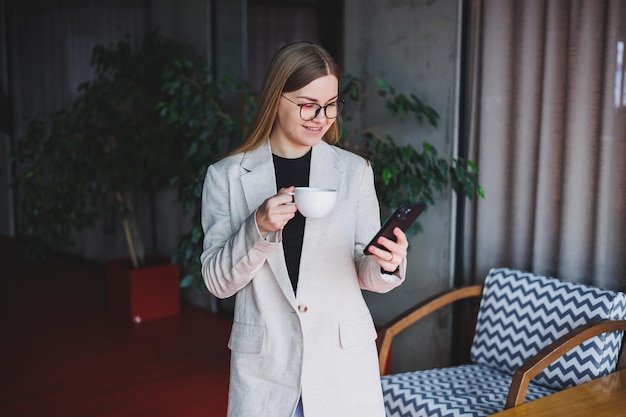 Pretty happy woman talking on a smartphone and drinking coffee\
while standing in a cafe young smiling blonde woman standing in a\
cafe concept of rest and free time modern female lifestyle