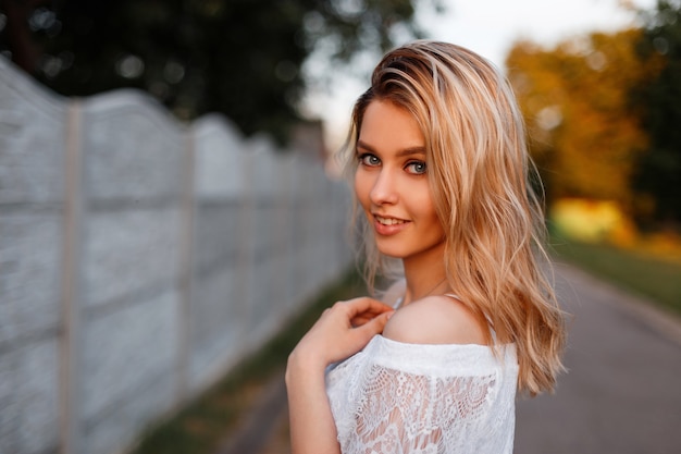 Giovane bella donna bionda abbastanza felice in un'elegante camicetta di pizzo bianco in posa all'aperto in una soleggiata giornata di primavera