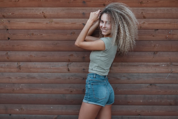 Pretty happy young stylish model woman with curly hairstyle in a t-shirt and trendy denim shorts stands near a wooden wall. Fun girl with smile enjoy the life