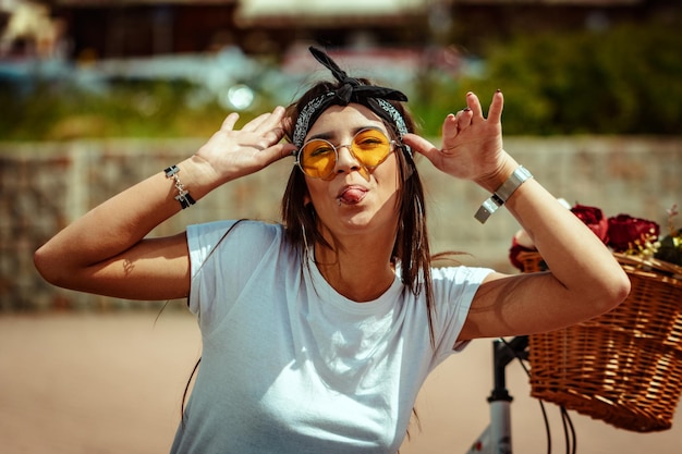 Pretty happy young woman is enyoing and having fun showing tongue, in a summer sunny day, beside the bike with flower basket.