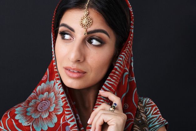 симпатичная индусская девушка в традиционном индийском платье сари и этнические украшения, покрывающая голову шарфом дупатта, изолированным над черной стеной