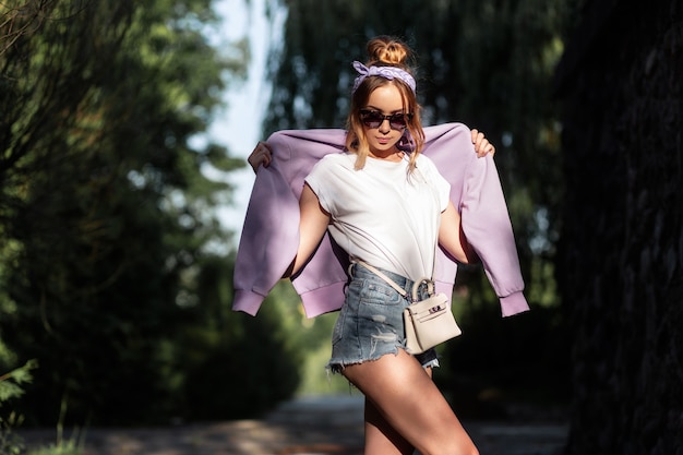 Ragazza graziosa hipster con occhiali da sole e una bandana in pantaloncini di jeans, una maglietta bianca e una felpa viola con una borsetta cammina all'aperto