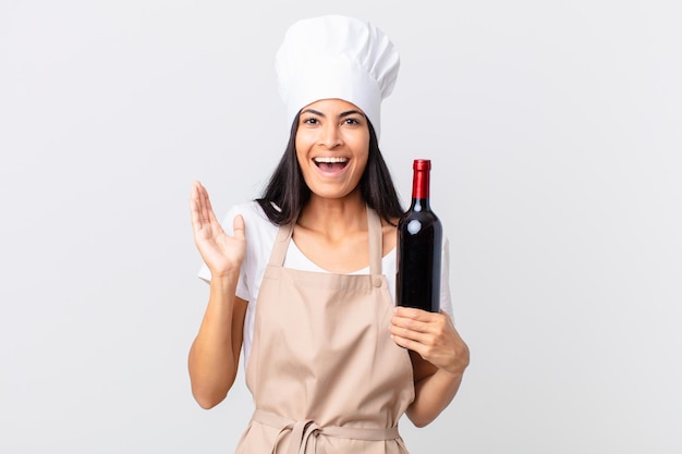 Pretty hispanic chef woman feeling happy and astonished at something unbelievable and holding a bottle of wine