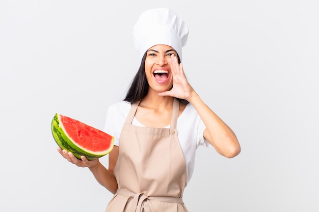 Pretty hispanic chef woman feeling happy,giving a big shout out with hands next to mouth and holding a watermelon