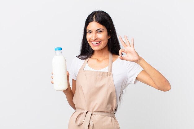 Pretty hispanic chef woman feeling happy, showing approval with okay gesture and holding a milk bottle