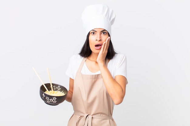 Pretty hispanic chef woman feeling shocked and scared and holding a noodle bowl