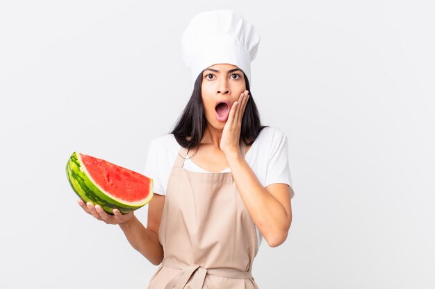 Pretty hispanic chef woman feeling shocked and scared and holding a watermelon