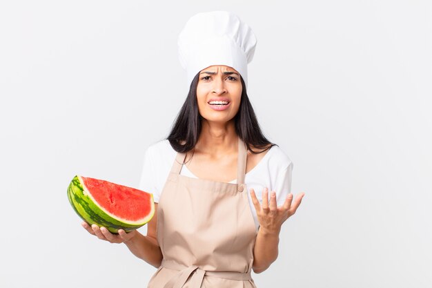 pretty hispanic chef woman looking desperate, frustrated and stressed and holding a watermelon