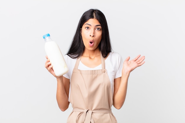 Pretty hispanic chef woman looking surprised and shocked, with jaw dropped holding an object and holding a milk bottle