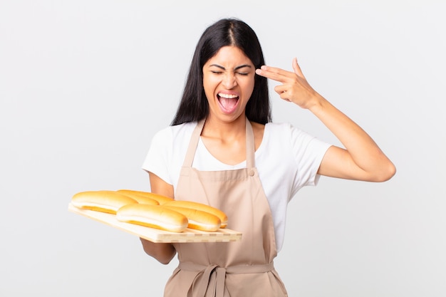 Pretty hispanic chef woman looking unhappy and stressed, suicide gesture making gun sign and holding a tray with bread buns