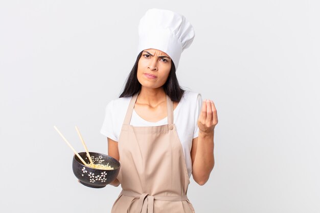 Pretty hispanic chef woman making capice or money gesture, telling you to pay and holding a noodle bowl