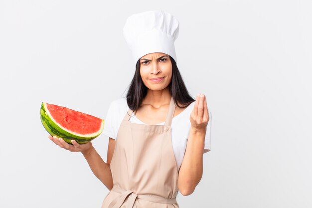 Pretty hispanic chef woman making capice or money gesture, telling you to pay and holding a watermelon