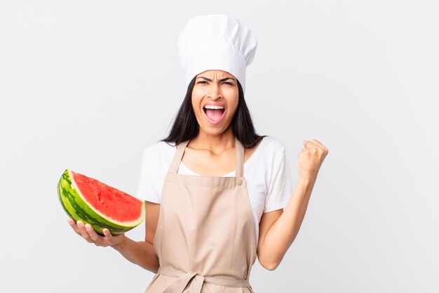 pretty hispanic chef woman shouting aggressively with an angry expression and holding a watermelon