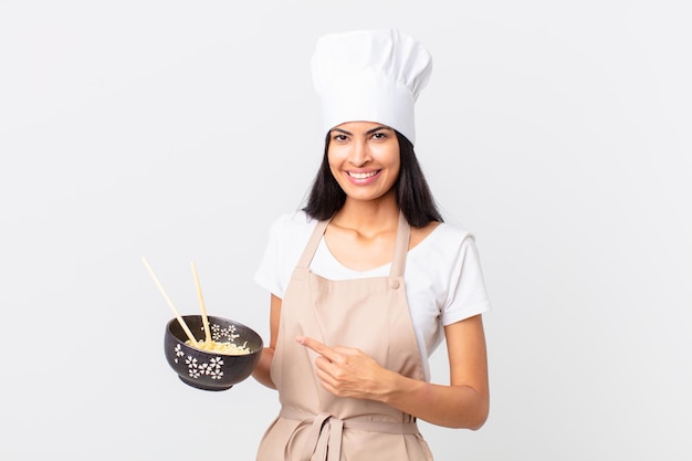 Pretty hispanic chef woman smiling cheerfully, feeling happy and pointing to the side and holding a noodle bowl