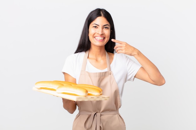 Bella donna ispanica chef sorridente che punta con fiducia al proprio ampio sorriso e tiene in mano un vassoio con focacce di pane