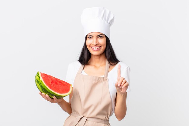 Pretty hispanic chef woman smiling and looking friendly, showing number one and holding a watermelon