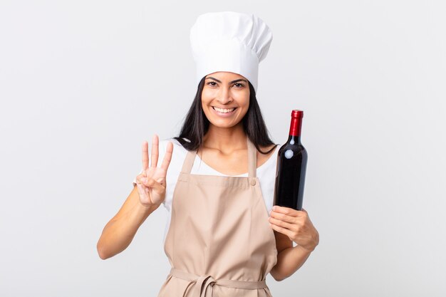 Pretty hispanic chef woman smiling and looking friendly, showing number three and holding a bottle of wine