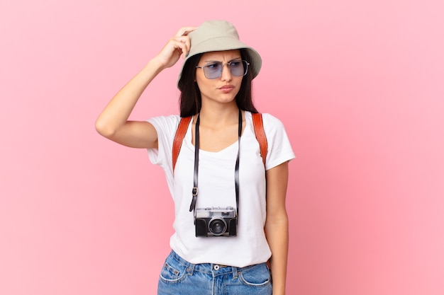 Pretty hispanic tourist feeling puzzled and confused, scratching head with a photo camera and a hat