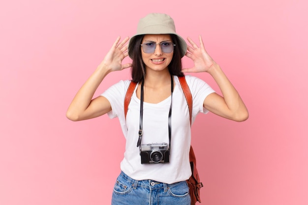 Photo pretty hispanic tourist feeling stressed, anxious or scared, with hands on head with a photo camera and a hat