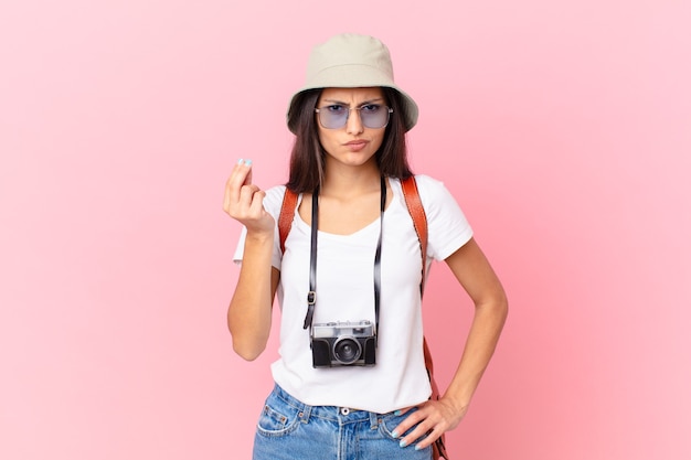 Photo pretty hispanic tourist making capice or money gesture, telling you to pay with a photo camera and a hat