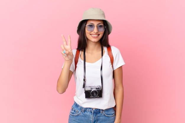 Pretty hispanic tourist smiling and looking friendly, showing number two with a photo camera and a hat