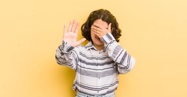 Pretty hispanic woman covering face with hand and putting other hand up front to stop camera refusing photos or pictures