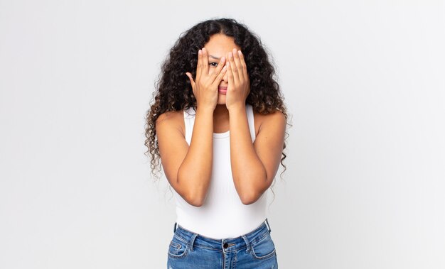 pretty hispanic woman covering face with hands, peeking between fingers with surprised expression and looking to the side