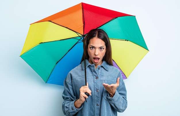Pretty hispanic woman feeling extremely shocked and surprised umbrella concept