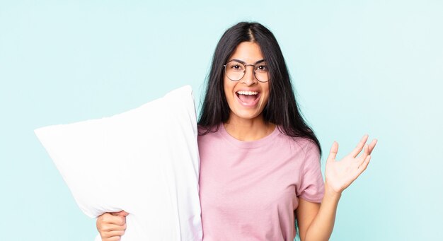 Pretty hispanic woman feeling happy and astonished at something unbelievable and wearing pajamas with a pillow