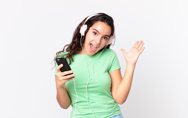 Pretty hispanic woman feeling happy and astonished at something unbelievable with headphones and a smartphone