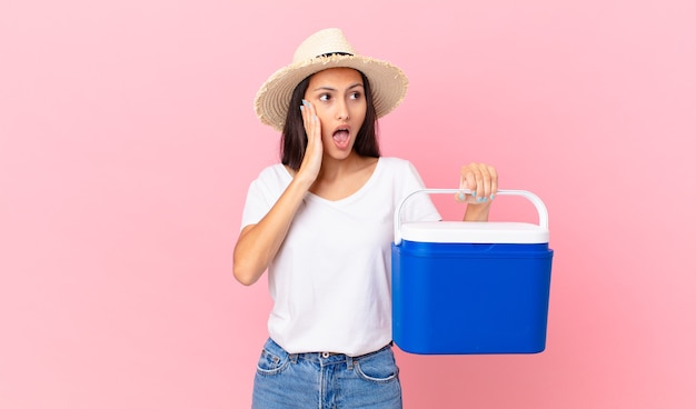 Pretty hispanic woman feeling happy, excited and surprised and holding a portable refrigerator