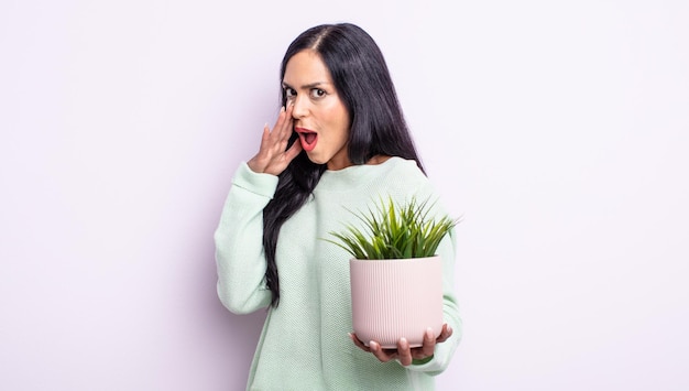 Pretty hispanic woman feeling happy,giving a big shout out with hands next to mouth. house plant concept