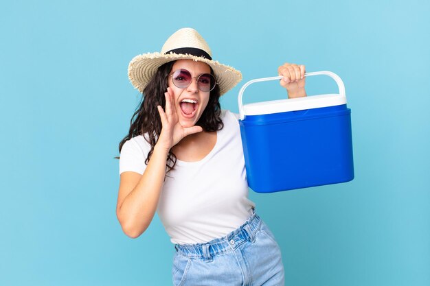 Pretty hispanic woman feeling happygiving a big shout out with hands next to mouth holding a portable refrigerator