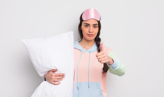 Pretty hispanic woman feeling proudsmiling positively with thumbs up pajamas and pillow concept