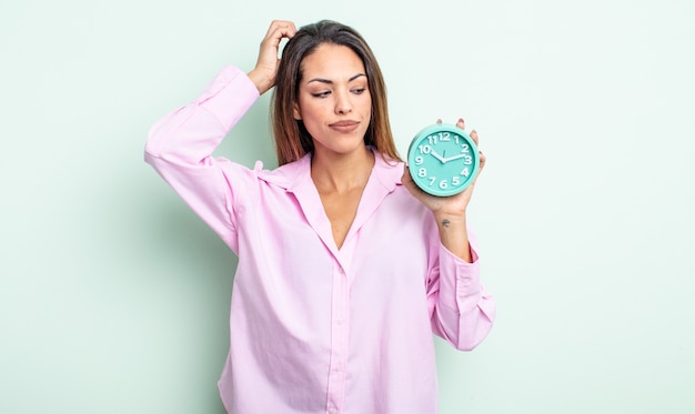 Pretty hispanic woman feeling puzzled and confused, scratching head. alarm clock concept