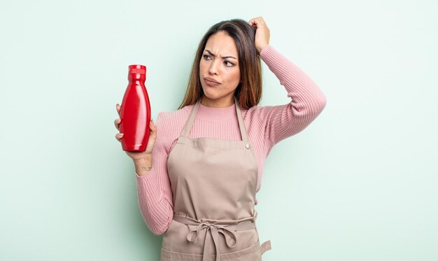 Pretty hispanic woman feeling puzzled and confused scratching head ketchup concept