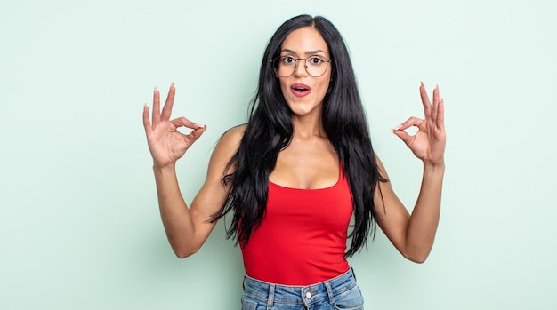 Pretty hispanic woman feeling shocked, amazed and surprised, showing approval making okay sign with both hands