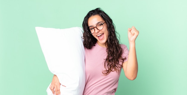 Pretty hispanic woman feeling shocked,laughing and celebrating success and wearing pajamas with a pillow