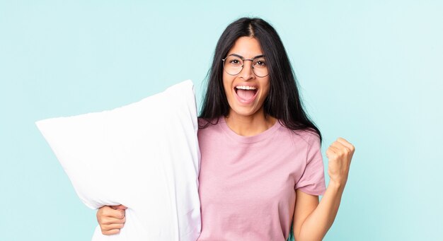 Pretty hispanic woman feeling shocked,laughing and celebrating success and wearing pajamas with a pillow