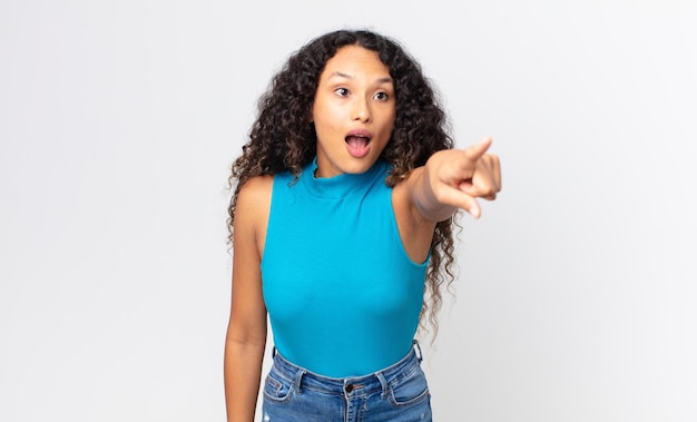 Pretty hispanic woman feeling shocked and surprised, pointing and looking upwards in awe with amazed, open-mouthed look