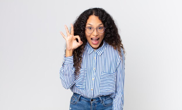 Pretty hispanic woman feeling successful and satisfied smiling with mouth wide open making okay sign with hand