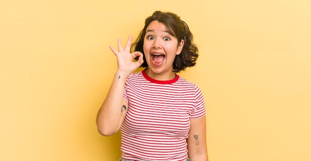 Pretty hispanic woman feeling successful and satisfied smiling with mouth wide open making okay sign with hand