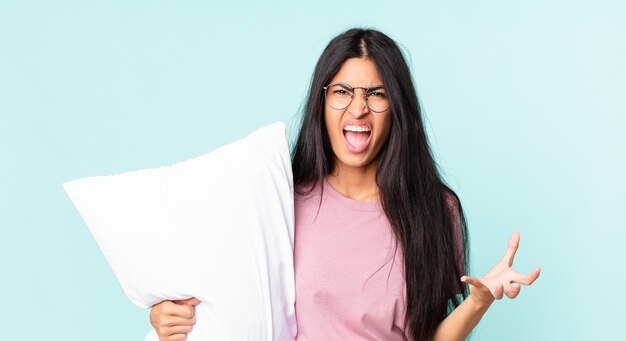 Pretty hispanic woman looking angry, annoyed and frustrated and wearing pajamas with a pillow