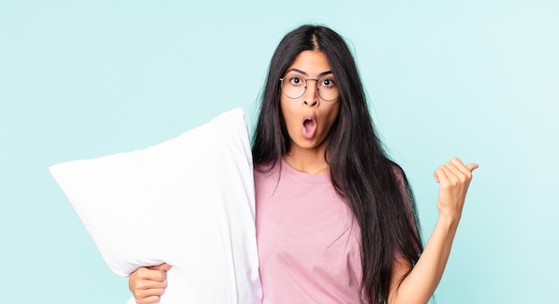 Pretty hispanic woman looking astonished in disbelief and wearing pajamas with a pillow