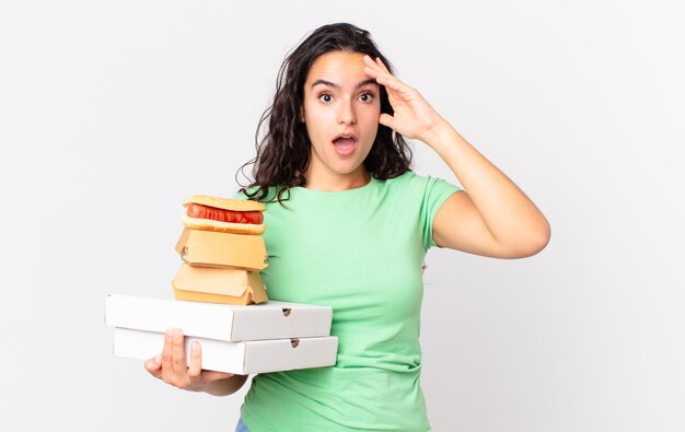 Pretty hispanic woman looking happy, astonished and surprised and holding take away fast food boxes