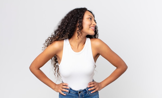 Pretty hispanic woman looking happy, cheerful and confident, smiling proudly and looking to side with both hands on hips