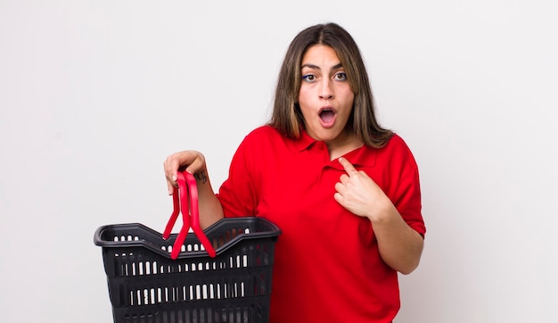 Pretty hispanic woman looking shocked and surprised with mouth wide open pointing to self empty shopping basket concept