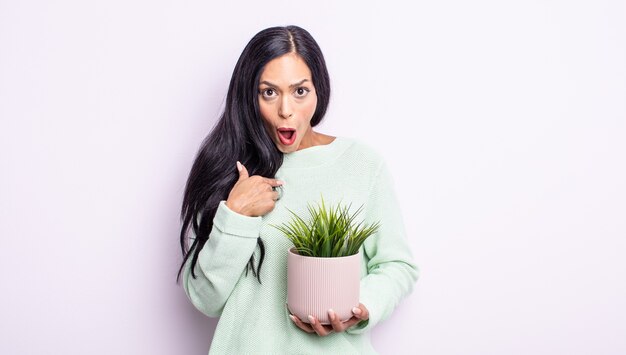 Pretty hispanic woman looking shocked and surprised with mouth wide open, pointing to self. house plant concept