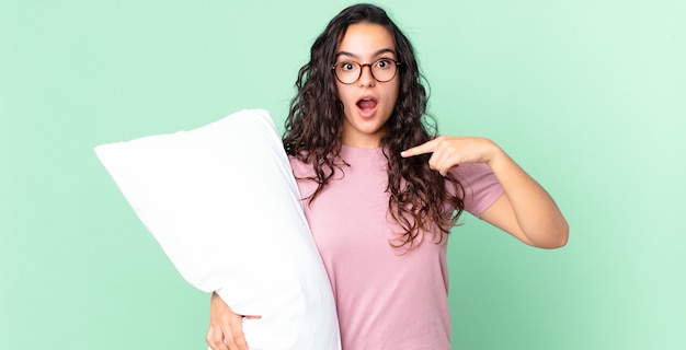 Pretty hispanic woman looking shocked and surprised with mouth wide open, pointing to self and wearing pajamas with a pillow